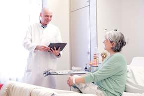 Chefarzt Dr. Dirk Mertens spricht mit Patientin die auf Krankenhausbett sitzt